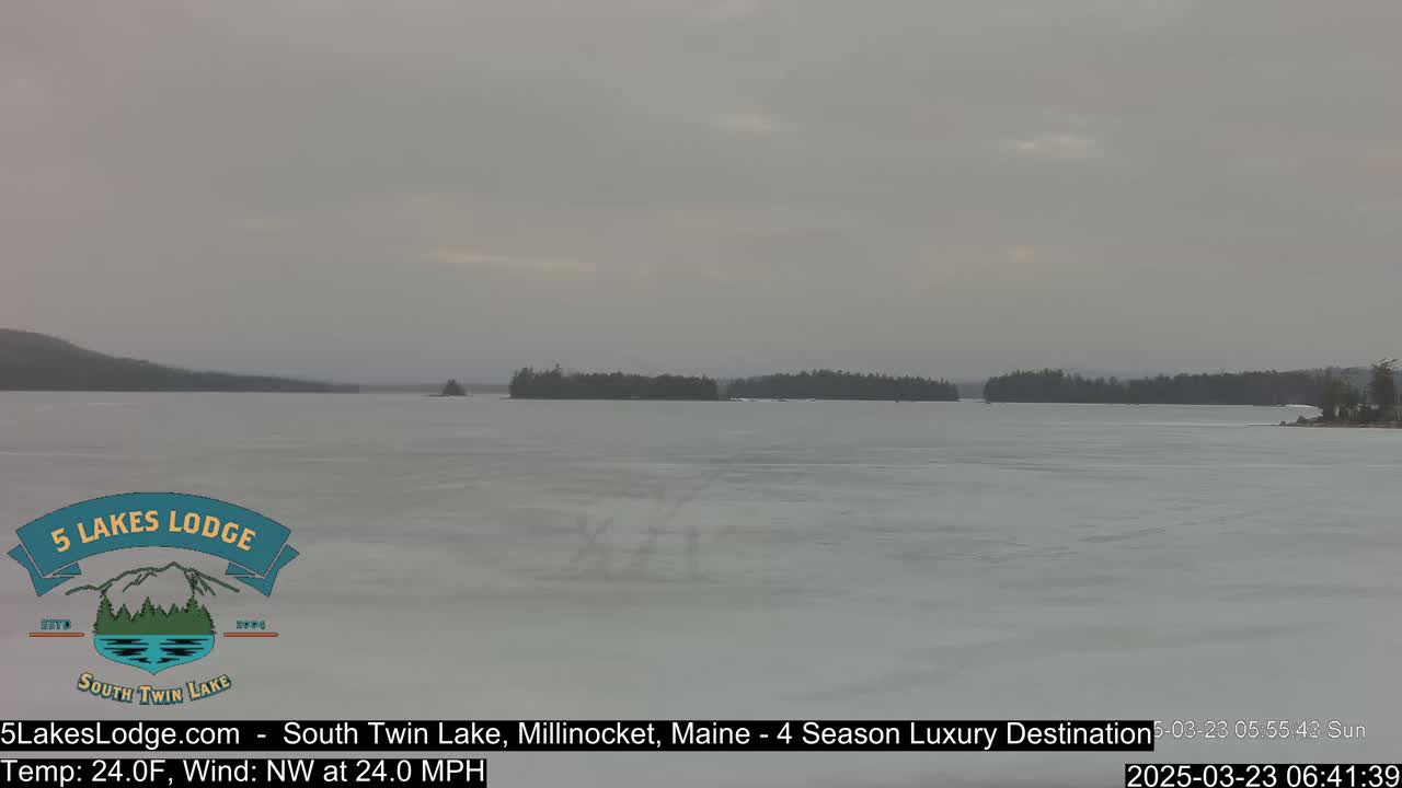 A mostly frozen lake is visible under an overcast sky, with a distant shoreline of dark trees.