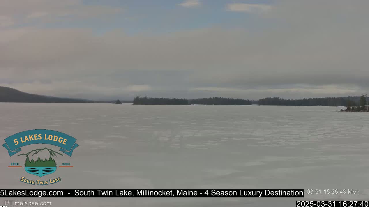A mostly frozen lake is visible under an overcast sky, with a treeline in the background.