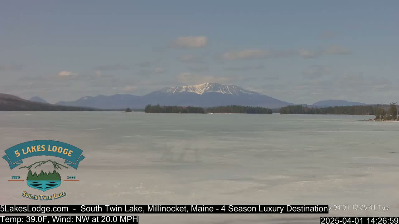 A partially frozen lake stretches to snow-capped mountains under a mostly clear sky.