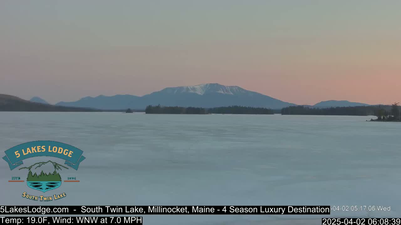 A partially frozen lake stretches towards a snow-capped mountain range under a pale pink sunset sky.