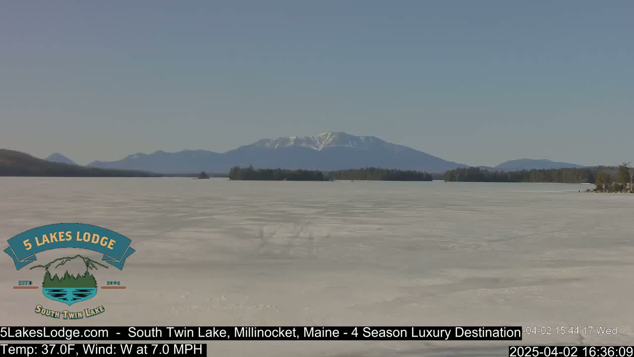 A mostly frozen lake stretches towards snow-capped mountains under a clear blue sky.