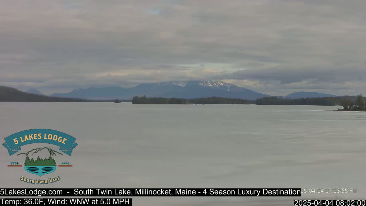 A mostly frozen lake stretches toward snow-capped mountains under a cloudy sky.