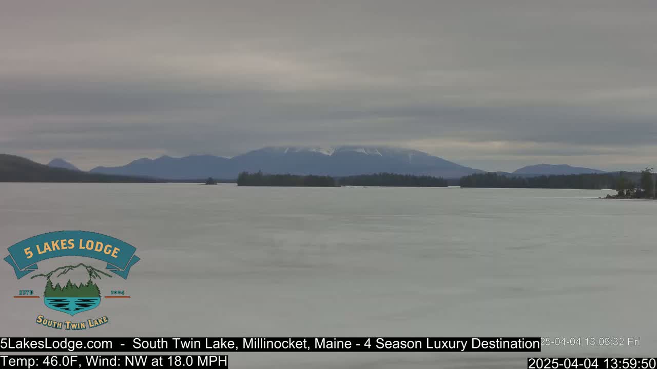 A partially frozen lake stretches toward snow-capped mountains under an overcast sky.