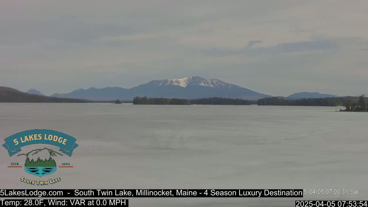 A partially frozen lake stretches toward a snow-capped mountain range under a cloudy sky.