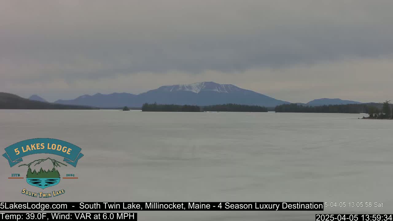 A partially frozen lake stretches out before a mountain range with a dusting of snow under an overcast sky.