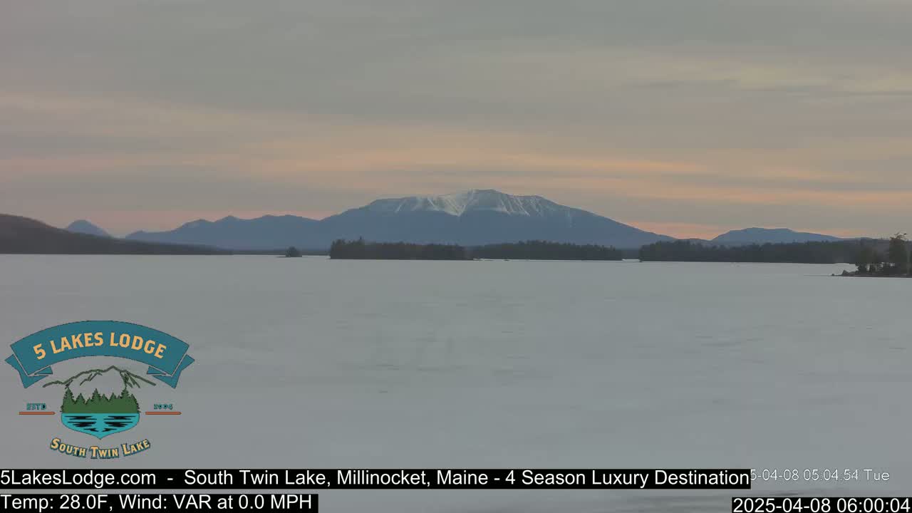 A snow-covered lake stretches toward a distant mountain range under a pale, dusky sky.