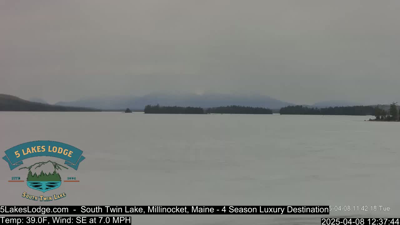 A mostly frozen lake is seen under an overcast sky, with a distant shoreline of dark trees and mountains.