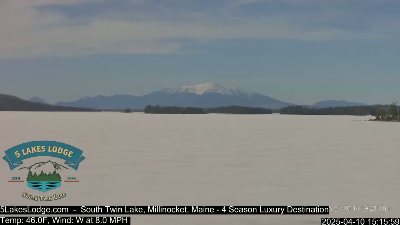 A snow-covered lake stretches to distant mountains under a partly cloudy sky.