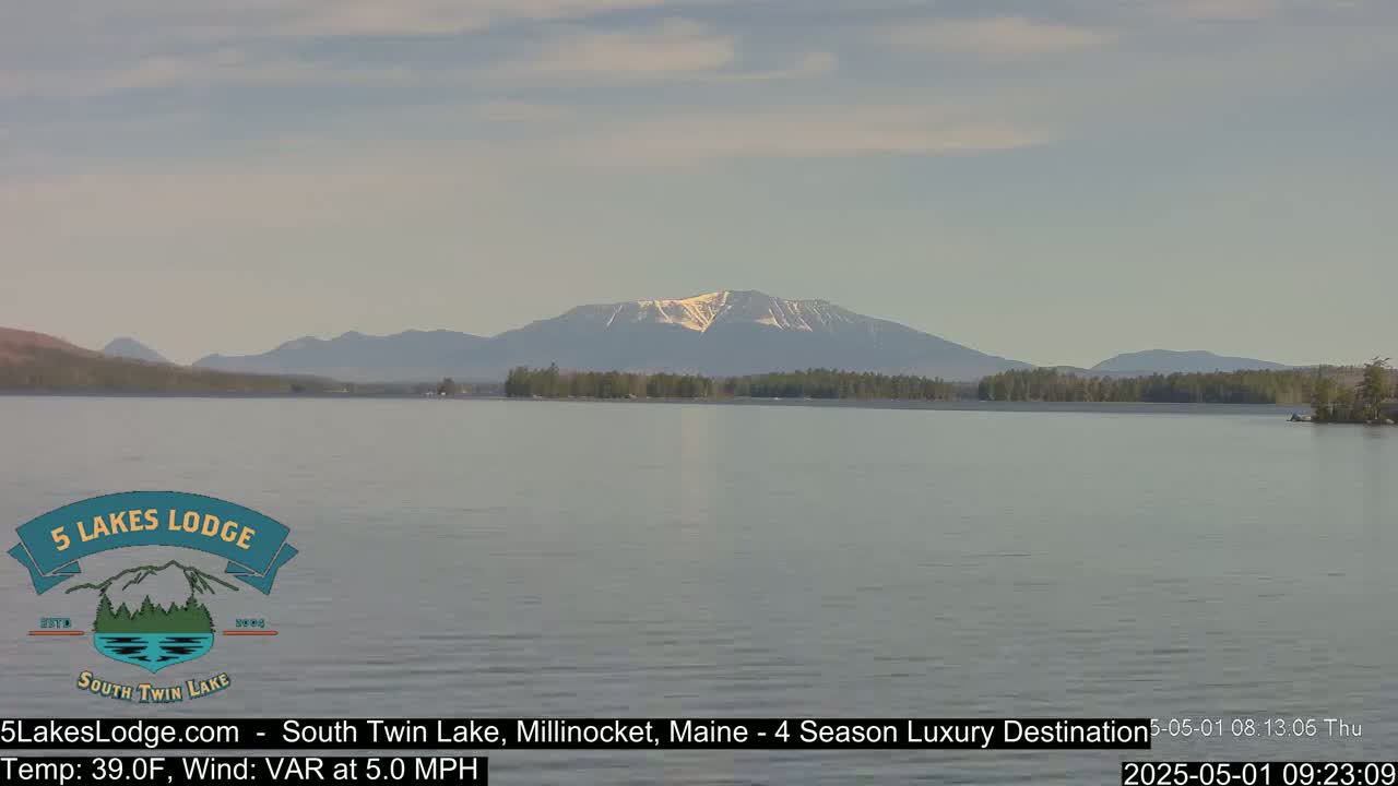 A calm lake with a mountain range in the background, partially covered in snow, under a mostly clear sky.