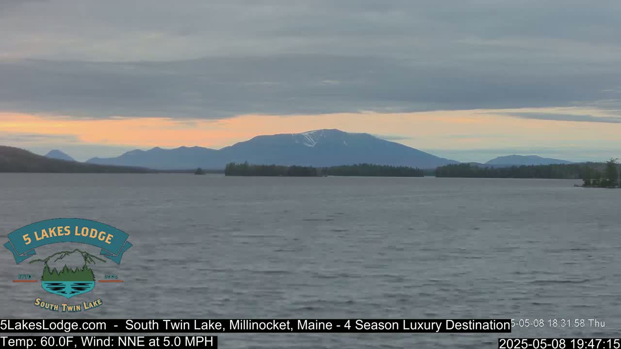 A calm lake with a mountain range in the background under a partly cloudy, pale orange and gray sky.