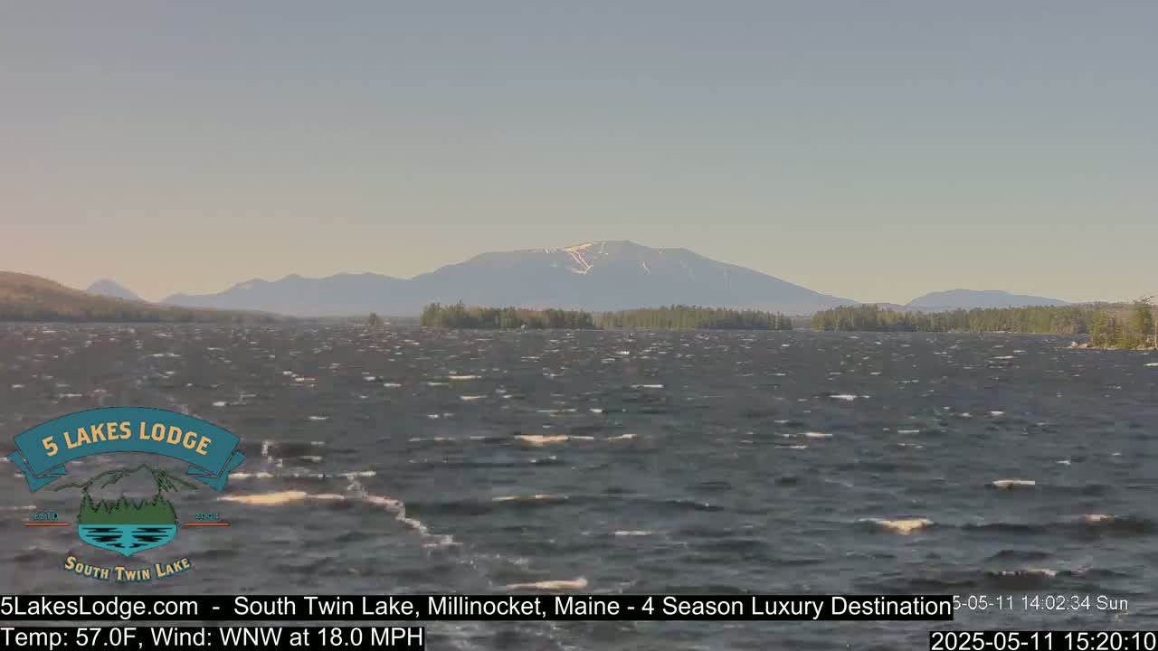 A windy lake with small waves stretches towards a mountain range under a mostly clear sky.