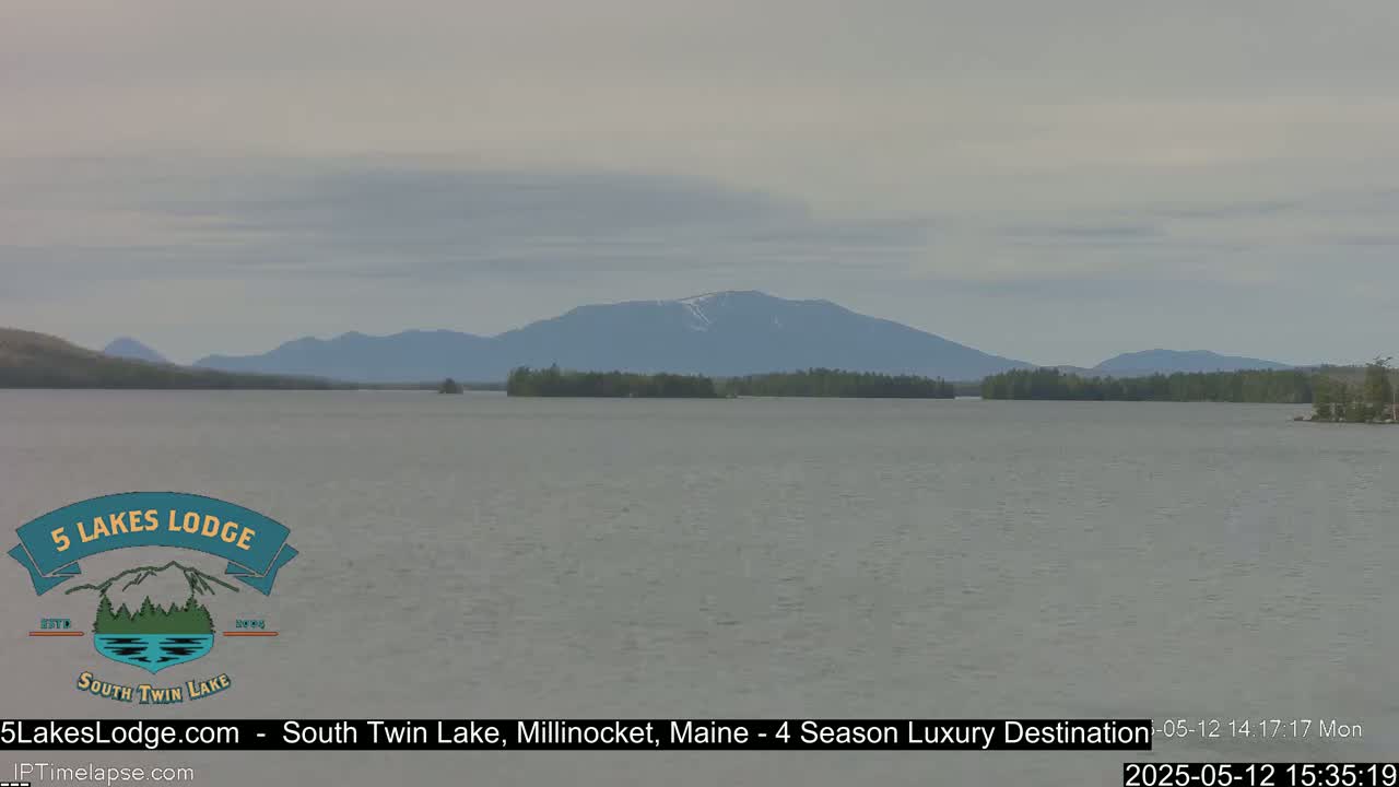A calm lake stretches out to distant mountains, one of which shows a patch of snow, under an overcast sky.
