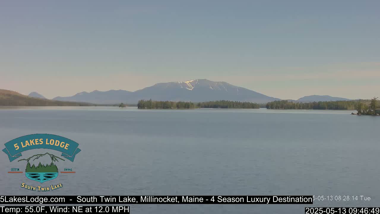 A calm lake stretches towards distant mountains, one of which has a patch of snow near its peak, under a clear, light-blue sky.