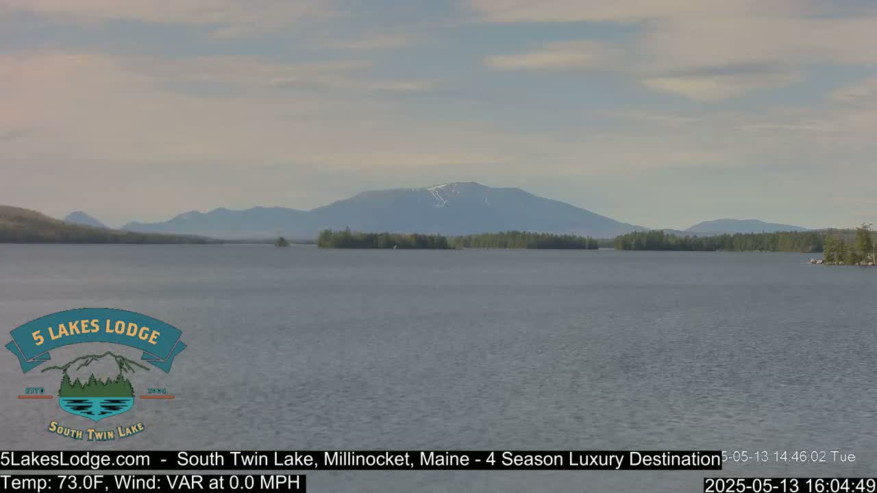 A calm lake is nestled among forested hills, with a distant mountain range partially covered in snow visible under a mostly cloudy sky.