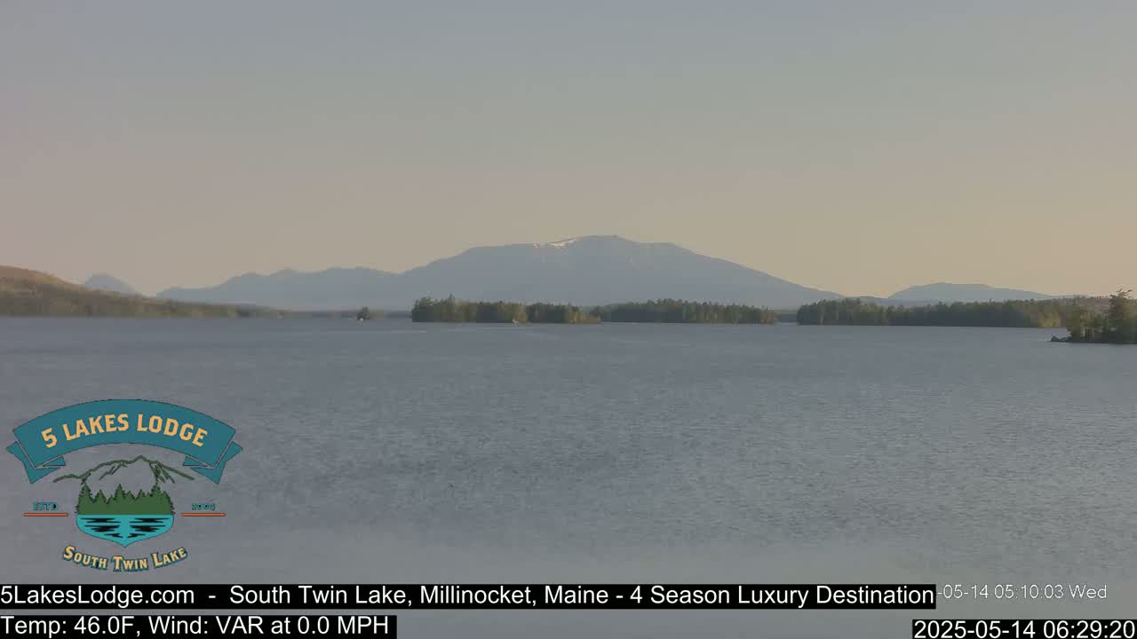 A calm lake stretches towards distant mountains, one of which has a patch of snow, under a clear, light-grey sky.