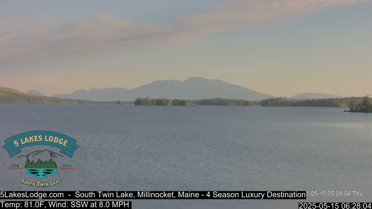 A calm lake stretches out to a mountain range under a partly cloudy, pale-blue sky.
