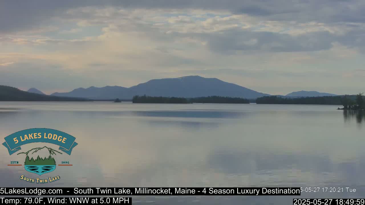 A calm lake reflects a mountain range under a partly cloudy sky.
