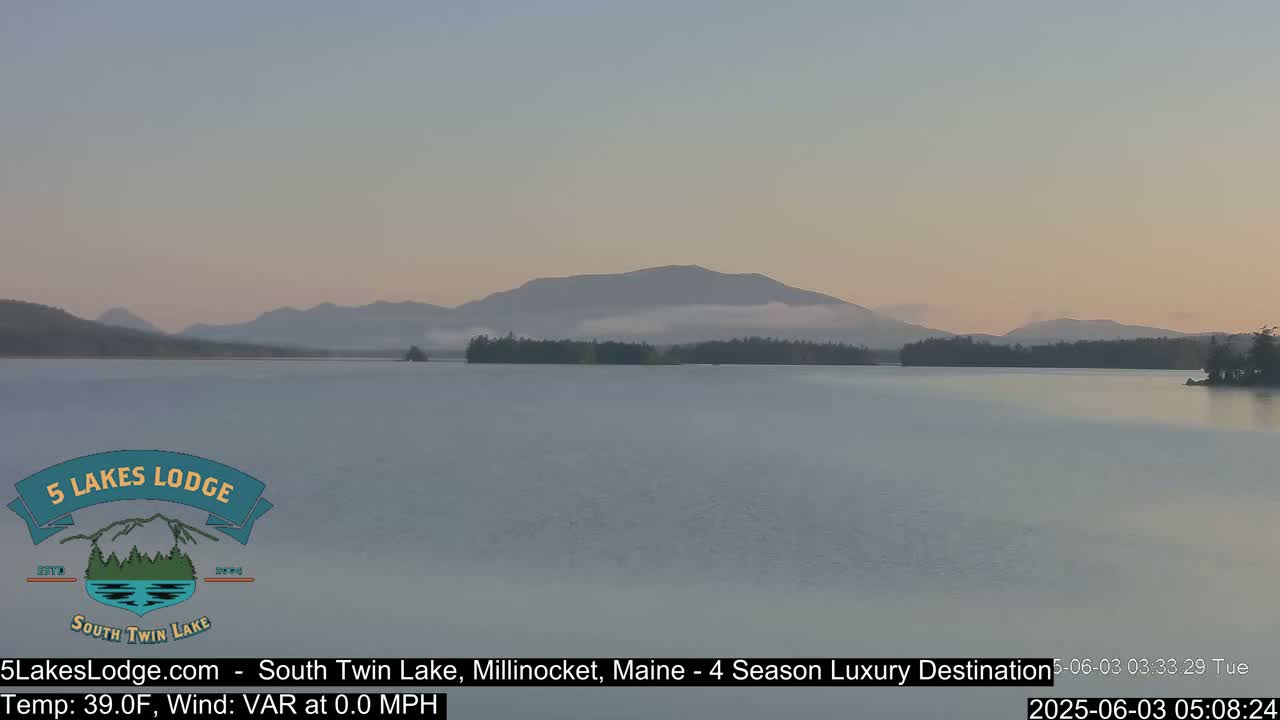 A calm lake at dawn, with mountains in the background partially shrouded in mist, under a pale sky.