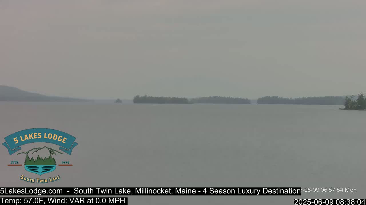 A hazy, calm lake is shown with a distant treeline under an overcast sky.