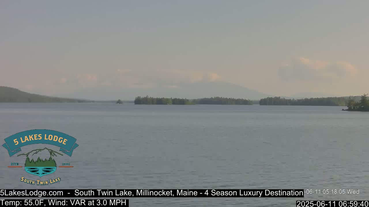 A calm lake under a mostly clear sky shows a distant treeline and mountains.