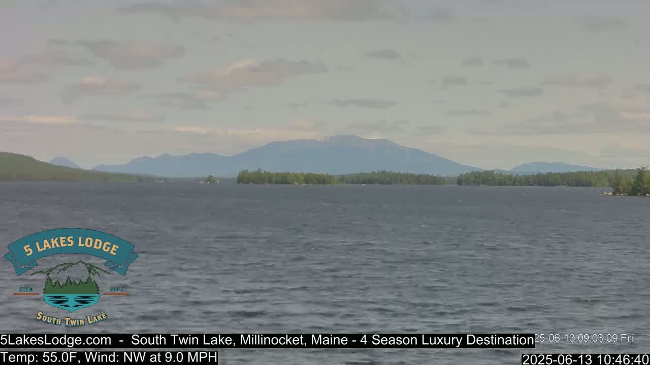 A lake with a gently rippling surface stretches toward distant mountains under a mostly cloudy sky.
