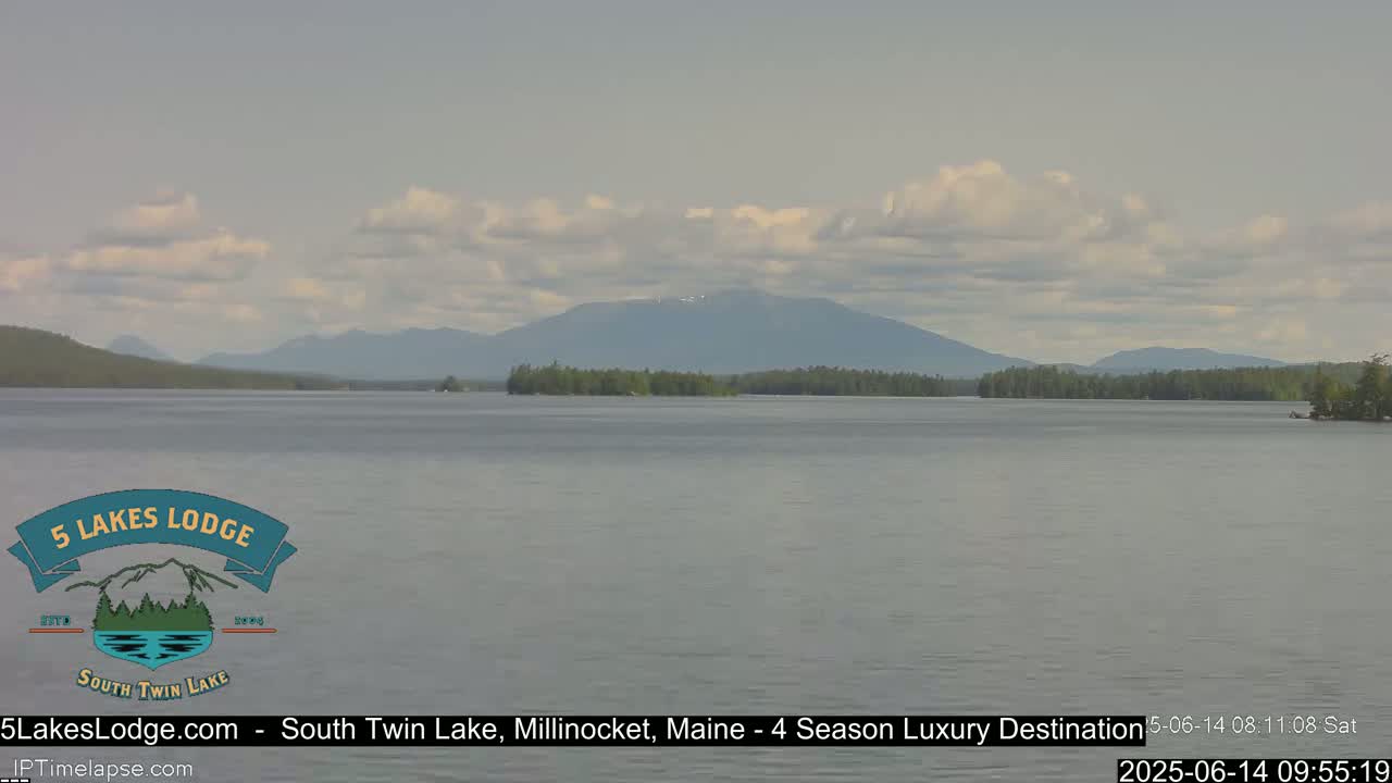 A calm lake stretches towards a mountain range under a partly cloudy sky.