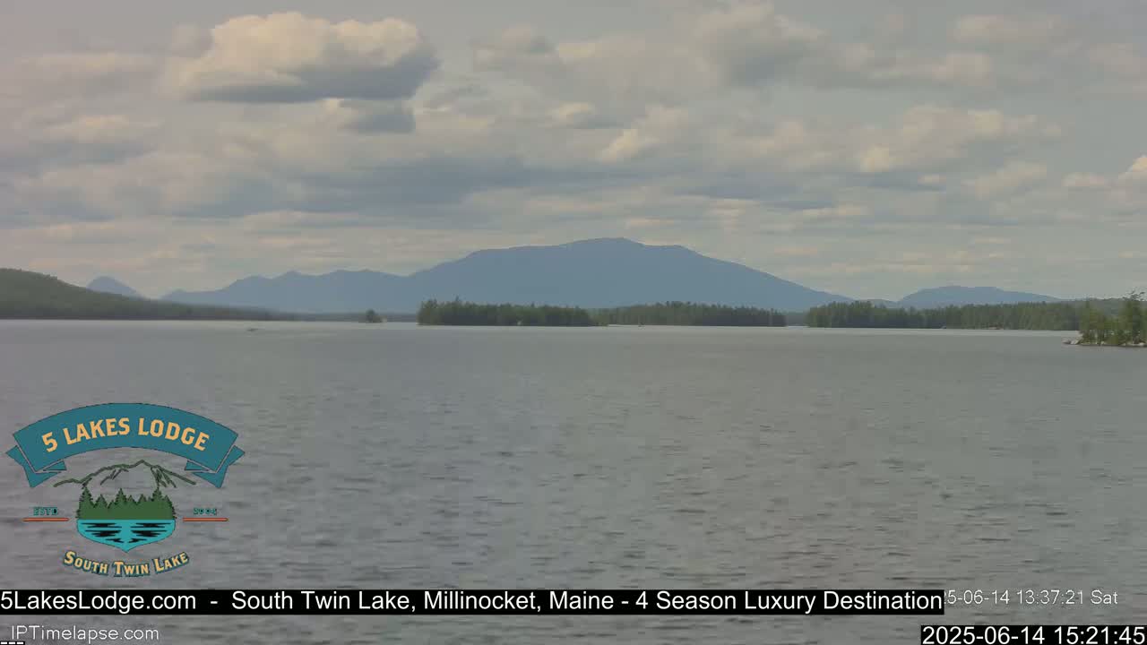 A calm lake stretches towards a mountain range under a partly cloudy sky.