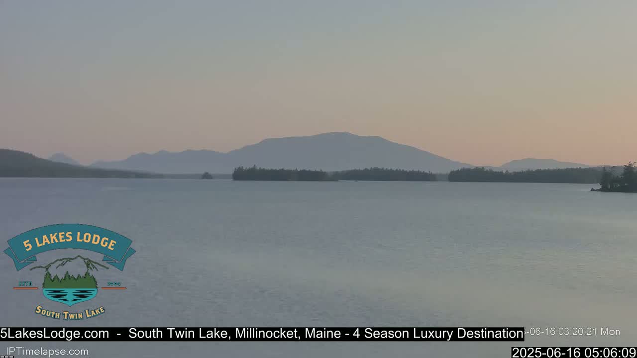 A calm lake stretches toward a hazy mountain range under a pale, pinkish sky.