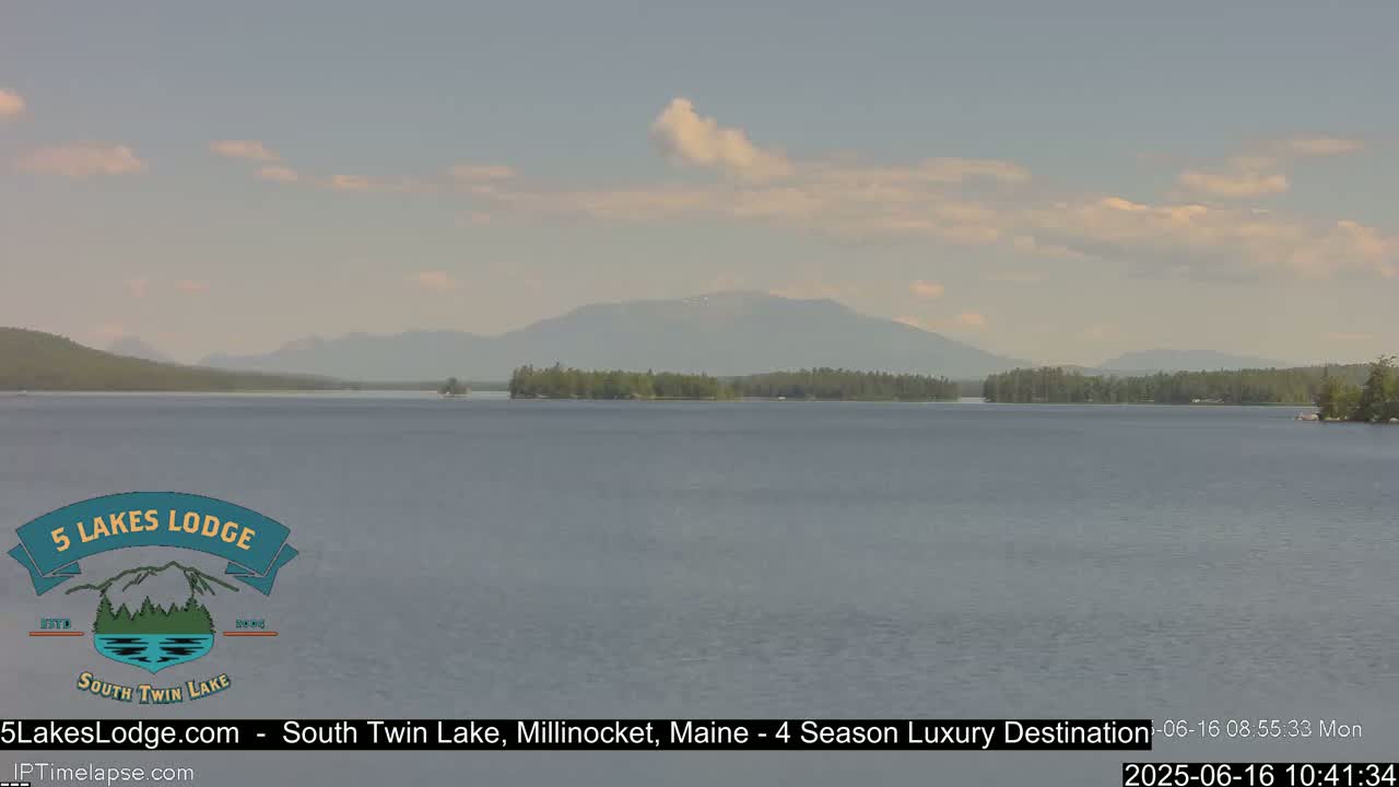 A calm lake stretches towards a mountain range under a partly cloudy sky.