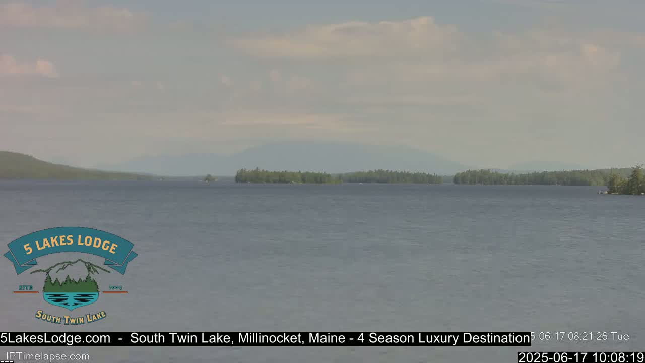 A calm lake with a distant tree-lined shoreline and mountains under a mostly cloudy sky.