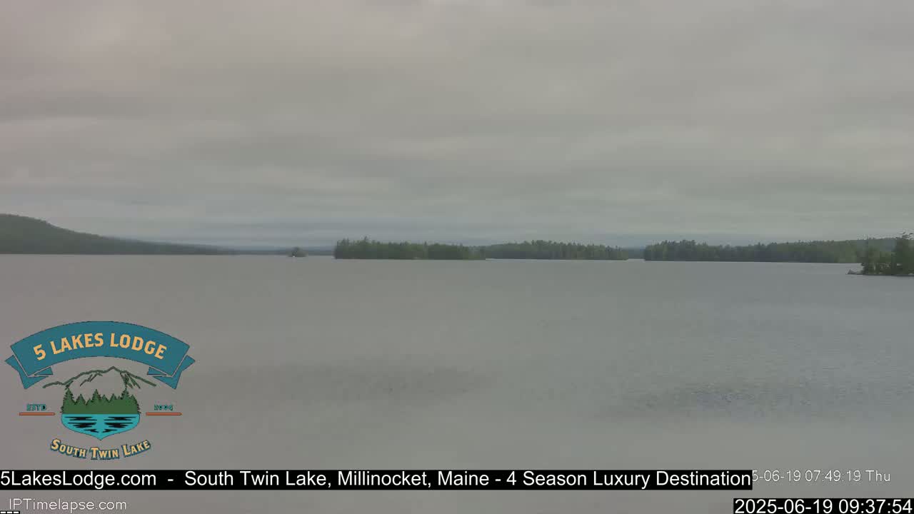 A calm lake is seen under an overcast sky, with a treeline visible in the distance.
