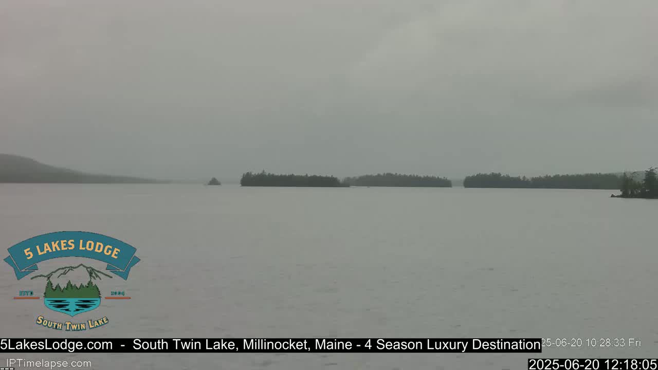 A calm lake under an overcast sky shows a distant shoreline of dark trees.