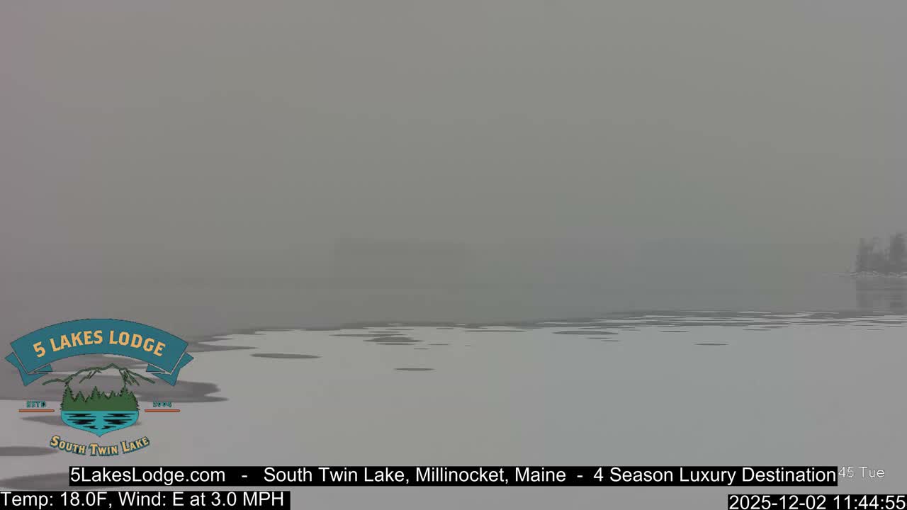 A partially frozen lake with patches of open water stretches under a uniformly gray, overcast sky, where low visibility creates a hazy horizon with only a faint outline of distant trees on the right.