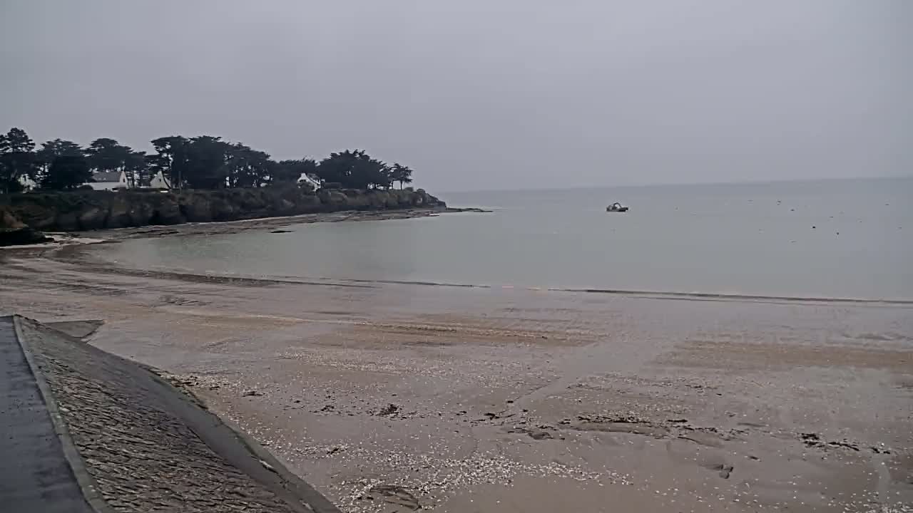 A calm, overcast day reveals a sandy beach at low tide, with a small boat on the pale water near a tree-lined headland.
