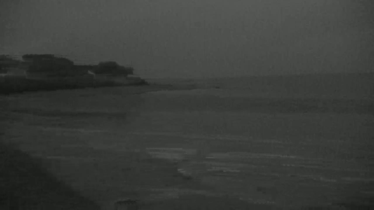 A dark, overcast sky hangs over a calm body of water lapping at a low, dark shoreline.