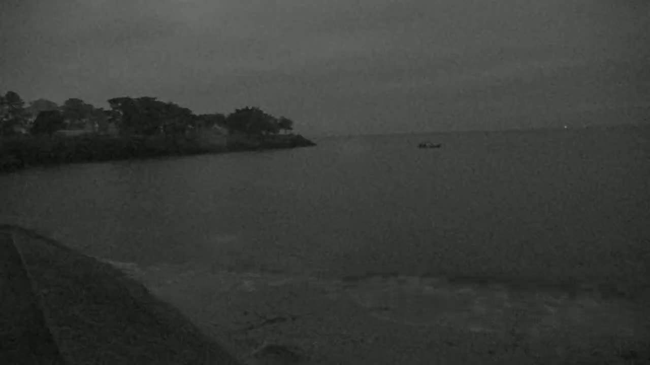 A dark, overcast day shows a calm body of water with a small cluster of trees and buildings on the shore, and a small boat in the distance.