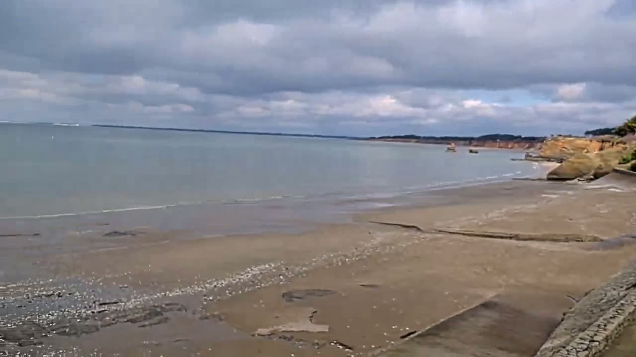 A sandy beach stretches out to a calm, light-grey sea under a mostly cloudy sky.