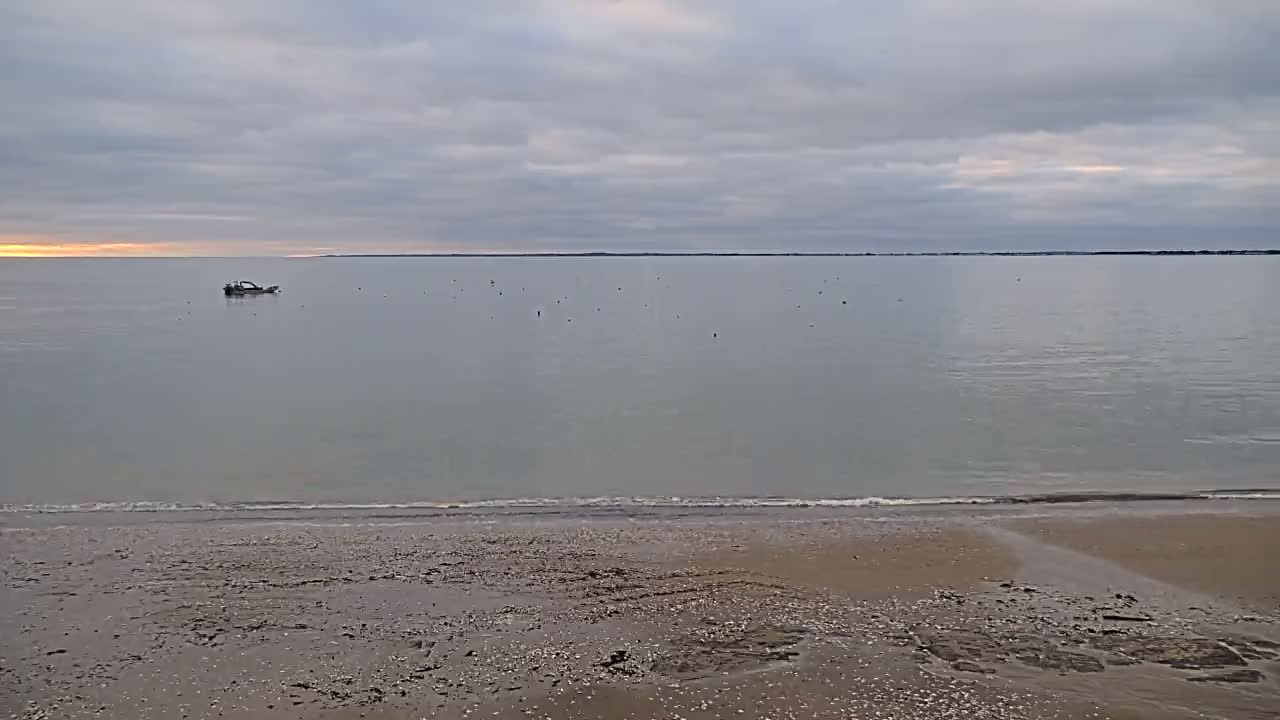 A small boat sits in calm, pale-grey water under a cloudy sky, on a beach with a gently sloping sandy shore.