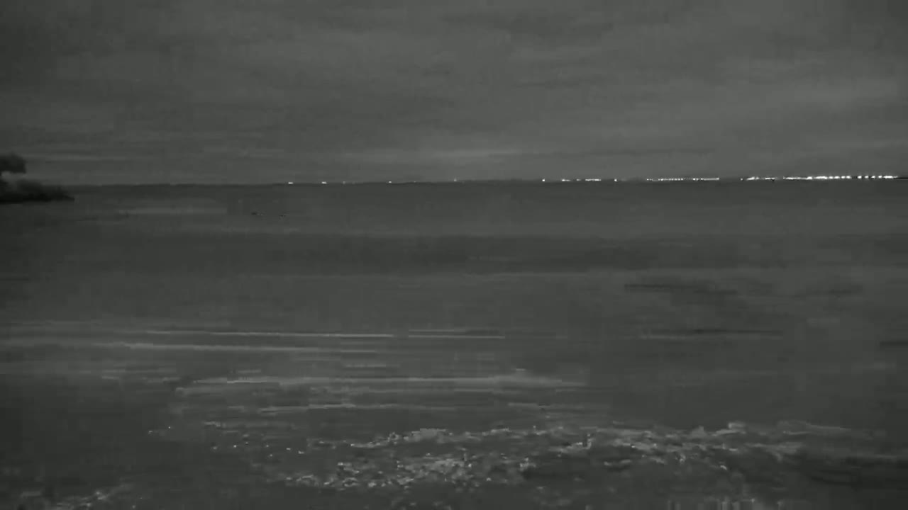 A dark, overcast night scene shows a body of water with a distant line of lights on the horizon.