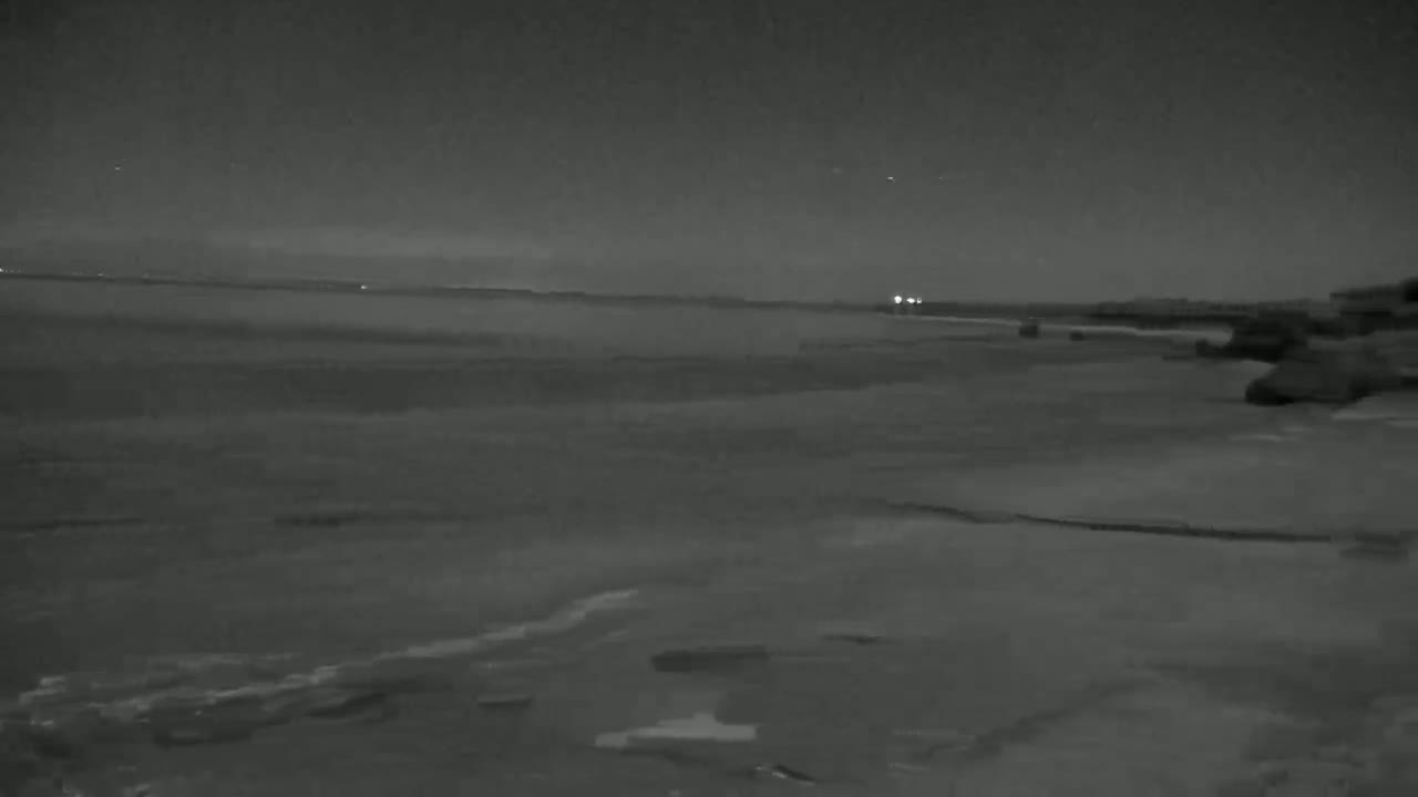 A dark, nighttime view of a calm body of water under a clear sky shows a shoreline with dark rocks and distant lights.