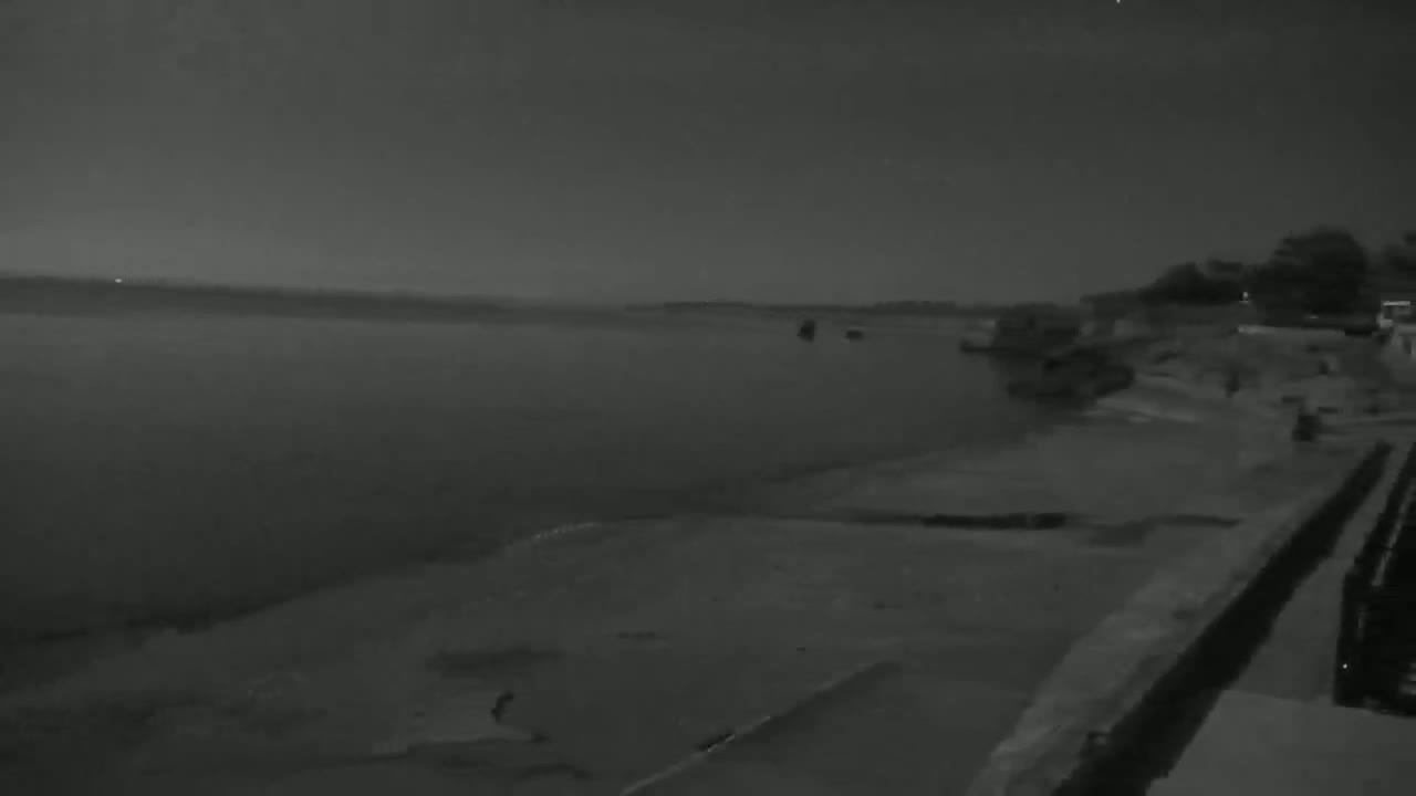 A dark, nighttime view shows a calm body of water stretching to a dark horizon under a clear sky, with a shoreline featuring some structures and vegetation.