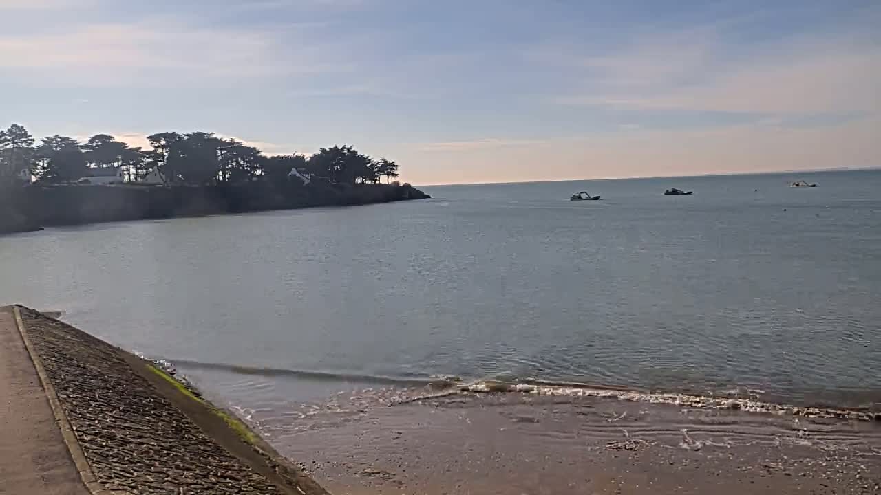A calm, light-grey sea laps a sandy beach below a rocky shoreline lined with dark green trees and houses, under a mostly clear sky.