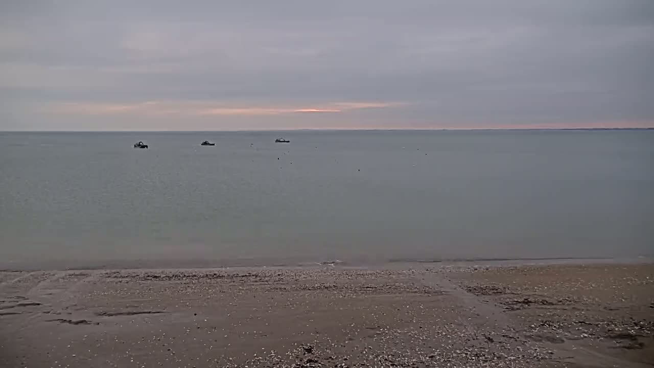 A calm, gray-sky day reveals a sandy beach meeting a flat, gray-green sea where several small boats are visible in the distance.