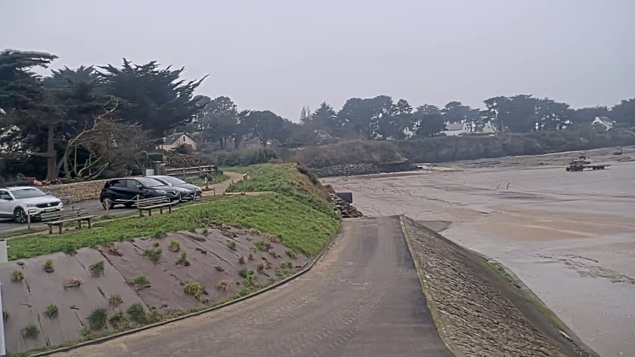 A paved boat ramp leads down to a wide expanse of exposed mudflats under an overcast sky, with several cars parked in a lot beside dark green trees and shrubs.