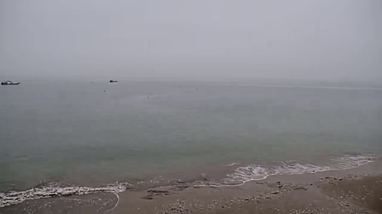 A hazy, overcast day shows a calm sea meeting a sandy beach with a few small boats visible in the distance.