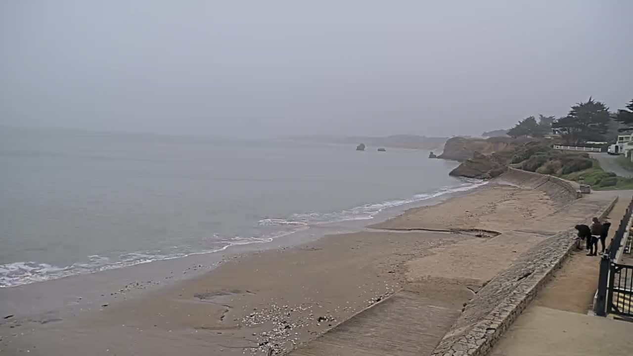 A foggy day at a sandy beach with gentle waves, a seawall, and a few people near some buildings in the distance.