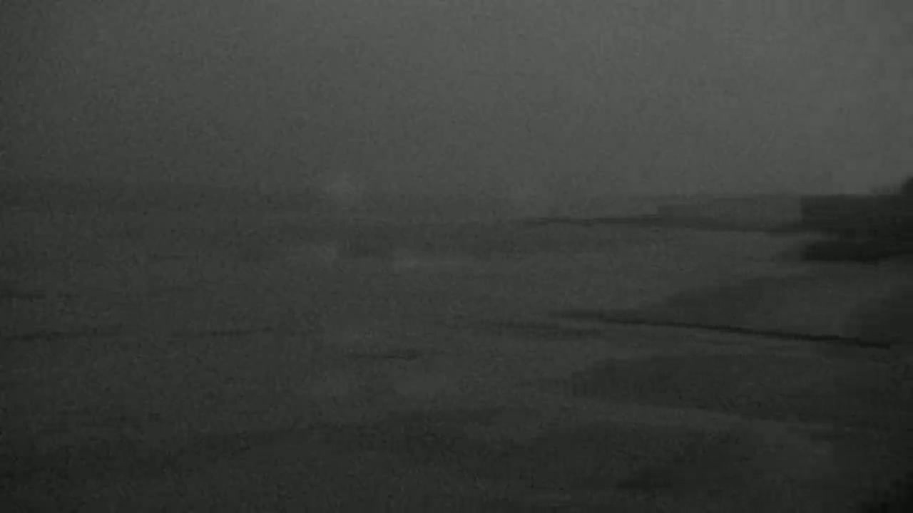 A hazy, dark gray scene shows a body of water and a dark shoreline under an overcast sky.