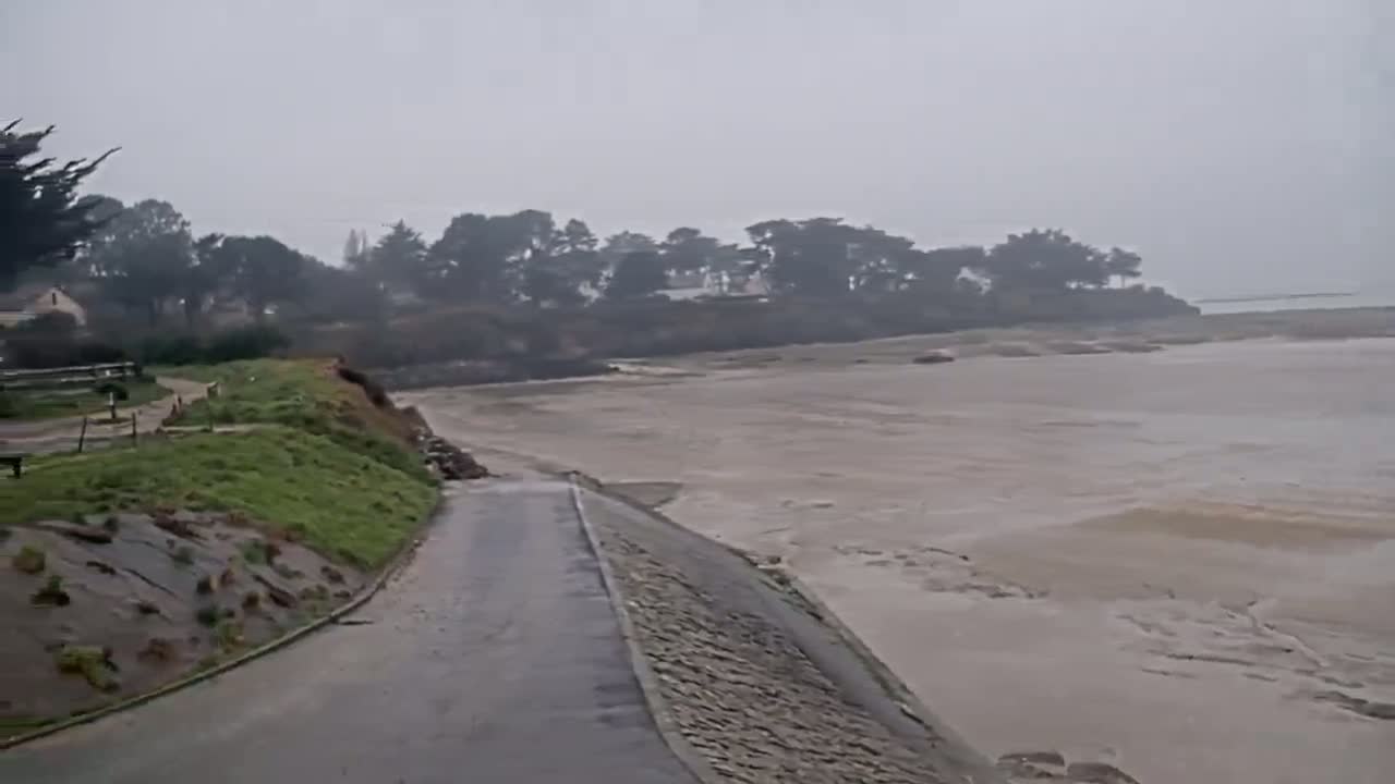 A paved road next to a grassy embankment overlooks a wide, muddy, and shallow body of water under an overcast sky.