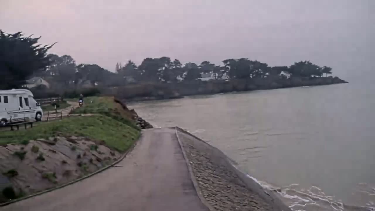 A white camper van is parked near a coastal road overlooking a calm, grey sea under an overcast sky.