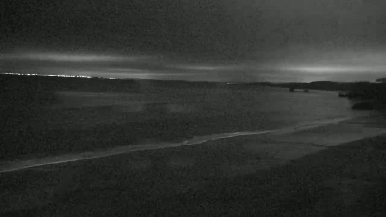 A dark, overcast night scene shows a calm body of water meeting a dark shoreline under a cloudy sky with distant lights visible on the horizon.